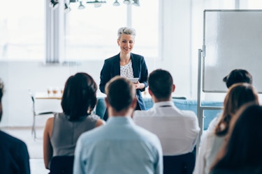  A smiling woman with short silver-blonde hair stands at the front of a conference room, facing an audience of employees sitting in black plastic chairs. A blank whiteboard stands to the right of the woman. 