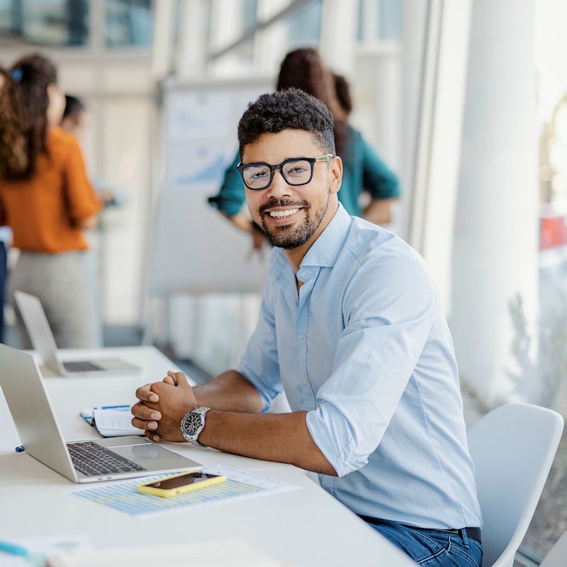 A young male office worker looks directly at the viewer and smiles. He is seated at a table with a laptop before him. Behind him in the background are office coworkers who are looking at a presentation easel.
