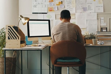  Rear view of a male entrepreneur working at a desk in his home office space. He has notes posted on the wall in front of him with to do lists, inspirational quotes, mock-up designs, and other notes. 
