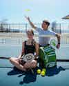 a man and woman sitting on a tennis court with rackets and balls