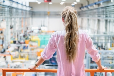 Back view of a woman business owner standing on a platform looking out onto the factory floor. 