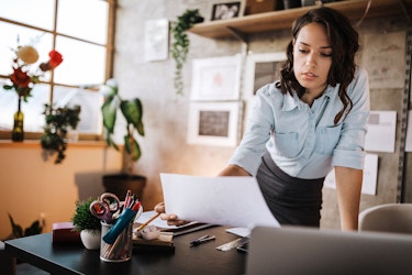  A business woman leans over her desk. She is holding up a paper in her left hand. She is reviewing an application as she prepares to finalize incorporation paperwork with her state government. 