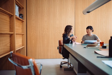  Two businesswomen are pictured in a modern office. They are sitting at a table and talking. 