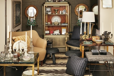  A Bergdorf Goodman display of Chairish's vintage furniture, laid out like a cluttered but elegant sitting room. The furniture includes two armchairs (one tan and one black with pinstripes), a pale gold shelving unit filled with various dishes and frames, several candelabras, three ottomans, a tall floor lamp, and two side tables (one close to the ground and one taller) laid with vintage dishes and glasses. The walls of the display are covered with mirrors and large paintings with ornate frames. 