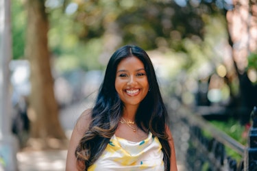  Cheyenne Sookoo, Founder of Paints by Chey, pictured from the chest up. Cheyenne has long dark hair and she wears a white sleeveless top with pops of teal and yellow on the fabric. She smiles and stands outside; the scenery behind her is artfully blurred and warped. 