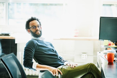  A young businessman leans back in an office chair with a slight smile on his face while he looks at the viewer. He is pictured in a modern office. Before him is a desktop computer and a mug. 