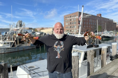  Mark Murrell, Founder and CEO of Get Maine Lobster, standing on a dock, holding a lobster in each hand, and making a "grrr" face for the camera.  Mark is a bald man with a thick white beard. He's wearing jeans and a black long-sleeved shirt with a multi-colored lobster printed on front. 
