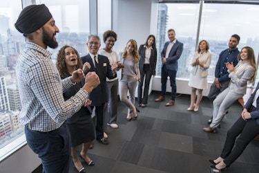  A group of about a dozen people in businesswear stand in a wide circle in an empty area of an office. The group's attention is on the man standing on the far left; he has a dark beard and wears a turbin and a windowpane plaid shirt. His fists are clenched in front of him in excitement and a couple of the other members of the group are applauding. The floor-to-ceiling windows behind the group show a sprawling cityscape. 