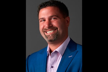  A smiling headshot of Jason Parker, Cofounder and CEO of K9 Resorts. Jason has dark hair and a goatee, and he is wearing a checked-print button-up shirt under a royal blue suit jacket. 