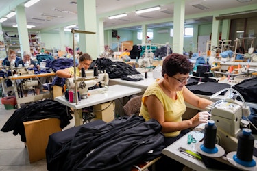  An angled shot of a line of women of various ages at sewing machines in a clothing manufacturing plant. Each woman has a pile of black or blue clothing next to her. 