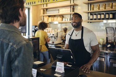  A man in a black apron stands behind a cash register in a coffee shop. A customer in a denim jacket stands across from him, facing away from the viewer. The cash register sits on a wooden counter and is made up of a large cash drawer and a mounted digital tablet. Behind the tablet, facing the customer, is a small sign reading "Exact Change Only." Behind the cashier, another employee, out of focus, works at a chrome coffee machine. 