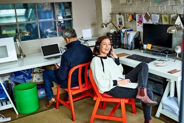  A woman small business owner sits on a red chair. She is talking on the phone with a customer and has a clipboard and notes on her lap to take notes during the call. 