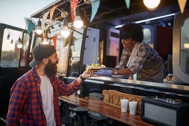  A woman food truck owner passes food to a male customer. 