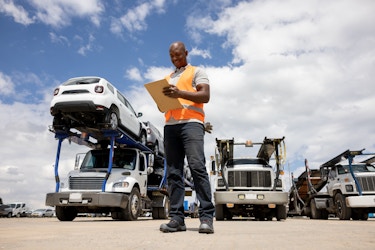  An employee stands in front of a fleet of trucks, looking down at the clipboard in his hands. The employee is bald and he wears a pale gray polo shirt, orange high-visibility vest, and black slacks. The trucks in the fleet are double-decker car carriers with white cabs. The truck on the far left is already loaded up with cars. 