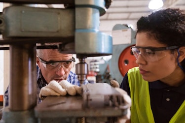  An older male instructor teaches a female apprentice to use factory machinery. 
